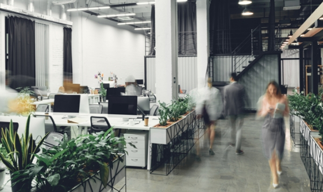 Modern open-plan office with white desks, computer monitors, and greenery, showing blurred motion of people walking through the space, with a staircase and industrial-style lighting in the background.