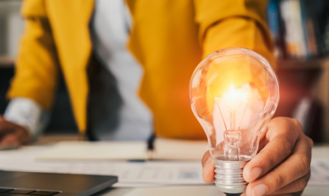 Person in a yellow blazer holding a glowing lightbulb at a desk with a laptop and papers, symbolizing an idea or innovation.